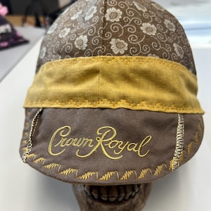 Pode incluir: Um boné castanho com um padrão floral na copa, uma faixa amarela mostarda sólida e uma aba castanha com o texto Crown Royal bordado em fio dourado. A aba é rematada com costuras decorativas em ziguezague douradas nas bordas.