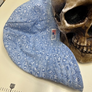 May include: A light blue paisley print welding cap with a white paisley design. The cap has a tag that says "Made in USA". A skull is in the background.