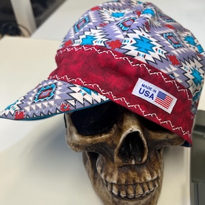 Puede incluir: Una gorra de soldadura con estampado rojo, blanco y azul, con una visera roja y una etiqueta "Made in USA". La gorra está sobre una calavera.