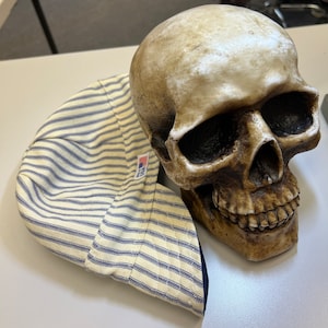 May include: A yellow and blue striped fabric hat with a blue brim and a small American flag patch. The hat is sitting on top of a white ceramic skull.