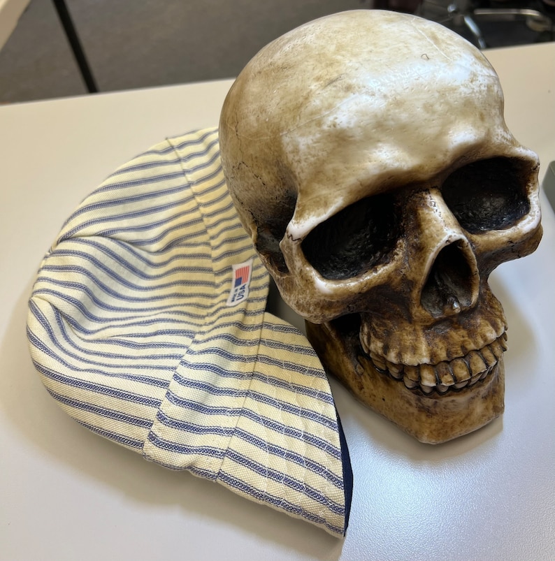 May include: A yellow and blue striped fabric hat with a blue brim and a small American flag patch. The hat is sitting on top of a white ceramic skull.