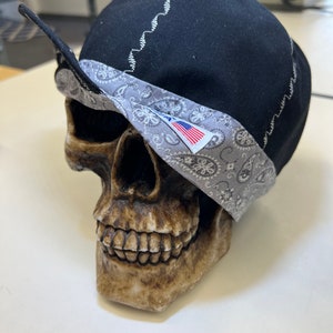 Op de afbeelding: Een zwarte laskap met een grijze paisley-patroonrand en witte stiksels. De kap wordt getoond op een schedelvormig decoratief object. De rand heeft een klein label met een Amerikaanse vlag.