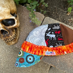 Op de afbeelding: Een veelkleurige laspet van stof met een paisley-patroon, een dierenprint uit het bos en een vlammenpatroon. De pet heeft een bruine stoffen rand en een label met de tekst "Made in USA".