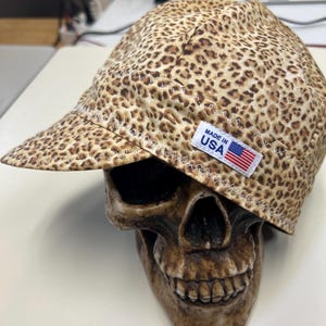 Pode incluir: Um boné com estampa de leopardo apresentando uma etiqueta retangular branca com o texto MADE IN USA e uma pequena bandeira americana. O boné é construído com costuras brancas em ziguezague visíveis ao longo da aba e das costuras.