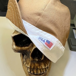 Op de afbeelding: Een bruine canvas werkmuts met een witte rand en een "Made in USA" label. De muts zit op een schedel.