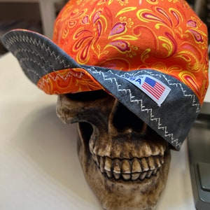 May include: A bright orange and yellow floral patterned baseball cap with a black brim and a small American flag patch. The cap is sitting on a skull.