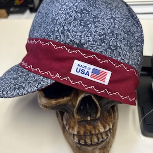 May include: A blue and white paisley patterned cap with a burgundy brim and white stitching. The cap has a small rectangular tag that reads "MADE IN USA" with an American flag. The cap is resting on a skull.