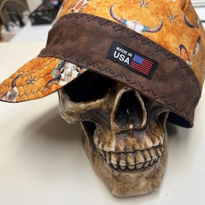 Pode incluir: Um boné de tecido com fundo laranja e um padrão de caveiras e estrelas. O boné tem uma faixa castanha larga com costuras em ziguezague e uma etiqueta retangular preta com a inscrição MADE IN USA e uma bandeira americana.