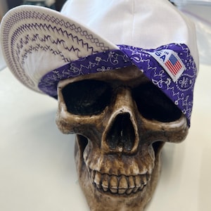Pode incluir: Um boné branco com uma faixa e aba paisley roxa, repousando sobre um crânio marrom. A aba tem costuras decorativas. Uma pequena etiqueta na faixa diz "Made in USA" com uma bandeira americana.