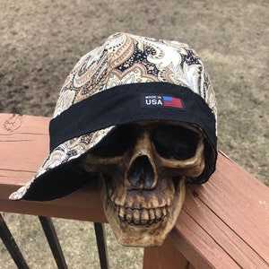 May include: A black and brown paisley patterned bandana style hat with a black brim and a black band around the crown. The band has a white label that says "Made in USA" with a small American flag.