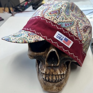 Puede incluir: Una gorra de soldador con un patrón de cachemir multicolor sobre fondo crema y una banda granate sólida en la parte delantera. Una etiqueta rectangular blanca con el texto MADE IN USA y un pequeño icono de bandera estadounidense está cosida en la banda.