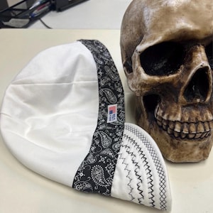 May include: A white welding cap with a black paisley patterned band and a black stitched edge. The cap has a small tag with a US flag. A brown skull is in the background.