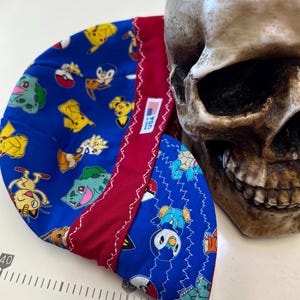 Pode incluir: Uma touca azul e vermelha estampada com vários personagens de desenhos animados. A touca é feita de tecido e tem uma costura branca. A etiqueta diz "Made in USA". Um crânio humano está no fundo.