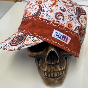 Könnte beinhalten: Eine Schweißerkappe mit weißem Stoffgrund und einem orangefarbenen, braunen und schwarzen Paisley-Muster. Die Kappe hat ein breites, rostfarbenes Band mit weißen Zickzacknähten und ein kleines weißes Etikett mit der Aufschrift MADE IN USA.