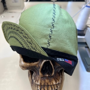 Puede incluir: Una gorra de béisbol verde y negra con un detalle de puntada en zigzag y una etiqueta "Made in USA".