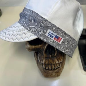 Può includere: Un berretto da saldatore bianco con una fascia a motivo paisley grigio e una visiera bianca con cuciture decorative. Una piccola etichetta rettangolare sulla fascia riporta il testo MADE IN USA accanto alla bandiera americana.