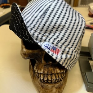 Może przedstawiać: Czarno-biała, bawełniana czapka w paski z czarnym skórzanym daszkiem i etykietą "Made in USA". Czapka leży na czaszce.