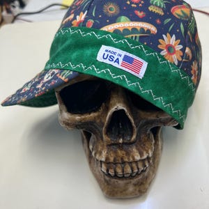 Puede incluir: Una gorra de soldador de tela azul oscuro con un patrón colorido de calaveras, sombreros y flores. La gorra tiene una banda verde ancha con costuras en zigzag blancas y una etiqueta rectangular blanca que dice MADE IN USA con una bandera.