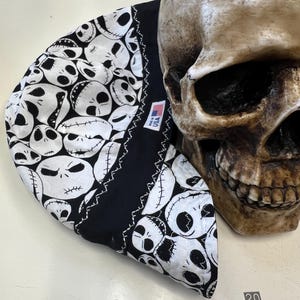 Può includere: Un berretto nero e bianco con un motivo ripetuto di facce di Jack Skellington. Il berretto ha un bordo nero con cuciture bianche e una piccola etichetta "USA". Un teschio decorativo è accanto al berretto.