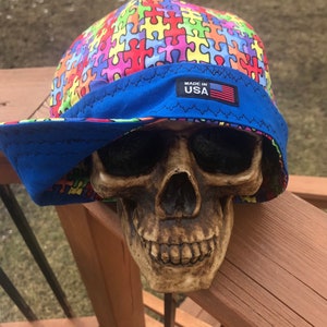 Könnte beinhalten: Ein blauer und mehrfarbiger Hut aus Stoff mit Puzzle-Muster, einem blauen Rand und einem "Made in USA"-Etikett. Der Hut sitzt auf einem Schädel.