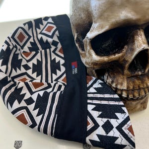 Könnte beinhalten: Schweißerkappe mit geometrischem Muster in Schwarz, Weiß und Braun. Die Kappe hat einen schwarzen Rand und ein Etikett mit der Aufschrift "MADE IN USA". Im Hintergrund ist ein Totenkopf zu sehen.