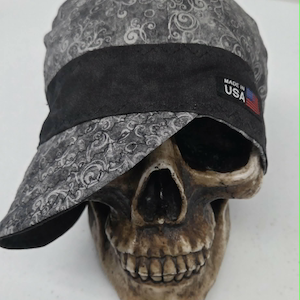 Pode incluir: Um boné de tecido cinza com um padrão paisley cinza escuro e uma faixa preta sólida na frente. Uma pequena etiqueta preta retangular na faixa exibe o texto branco MADE IN USA com uma pequena bandeira americana à direita.