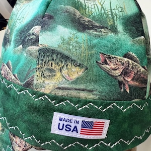 Op de afbeelding: Groene en blauwe werkmuts met een vismotief. Het heeft een wit label met de tekst "Made in USA" en een kleine Amerikaanse vlag.