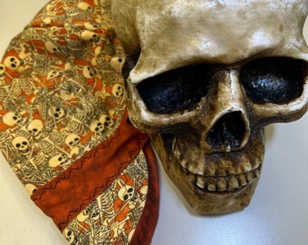 May include: A tan skull prop with dark eye sockets and a detailed jawline. A patterned fabric, featuring a repeating skeleton design in cream and black on an orange background, is draped over the skull.