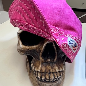 Puede incluir: Un sombrero de estilo bandana rosa con un patrón paisley blanco y las palabras "Made in USA" en el lateral. El sombrero está sentado sobre una figura de calavera.