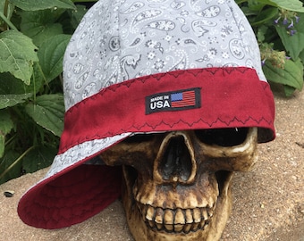 May include: A gray and white paisley bandana style welding cap with a red brim and a "Made in USA" label. The cap is sitting on a skull.