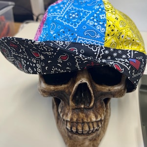 Op de afbeelding: Een zwarte, blauwe, roze en gele bandana-achtige hoed met een paisley-patroon. De hoed zit op een schedelfiguur.