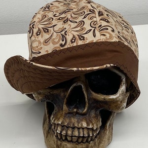 Peut inclure: Casquette de soudeur à imprimé cachemire marron et beige avec un bord marron foncé. Le chapeau est posé sur un crâne. La casquette a un bord incurvé et est en tissu. Le motif cachemire comprend des nuances de marron et de beige.