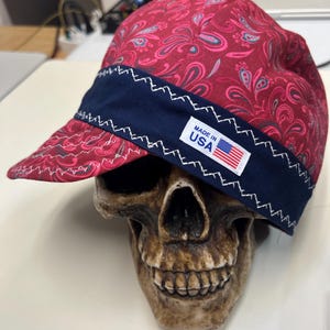 Pode incluir: Um boné vermelho com padrão paisley em rosa escuro e azul-petróleo, apresentando uma faixa azul-marinho na aba com costura em ziguezague branca. Uma etiqueta retangular branca traz o texto MADE IN USA e um pequeno ícone da bandeira americana.