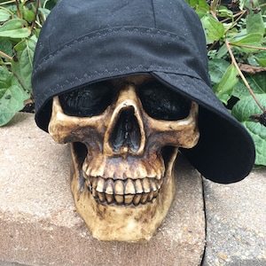 May include: A black fabric skull cap with a stitched brim sits on top of a white plastic skull. The skull has a detailed face with teeth and eye sockets.