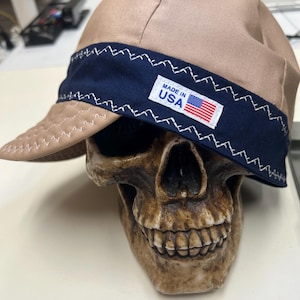 May include: A tan welding cap featuring a dark blue band with white zigzag stitching. A white rectangular label is sewn onto the band, displaying the text "MADE IN USA" alongside a small graphic of the American flag. The cap is placed on a skull.