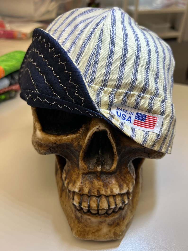 May include: A white and blue striped fabric hat with a navy blue brim and a "Made in USA" label. The hat is sitting on a skull.