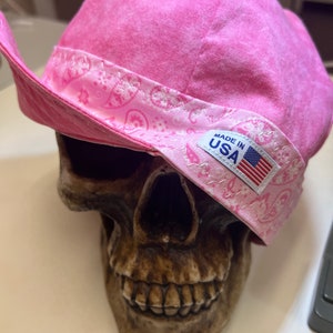 May include: A pink bandana style hat with a paisley pattern and a white "Made in USA" label. The hat is sitting on top of a skull.
