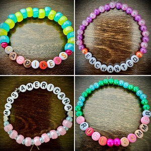 May include: Four colorful beaded bracelets with different words spelled out on them. The bracelets are: "Happiness", "Enchanted", "Miss Americana", and "Cruel Summer".