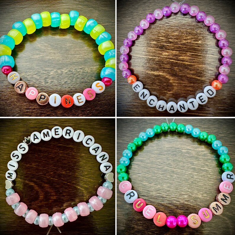 May include: Four colorful beaded bracelets with different words spelled out on them. The bracelets are: "Happiness", "Enchanted", "Miss Americana", and "Cruel Summer".