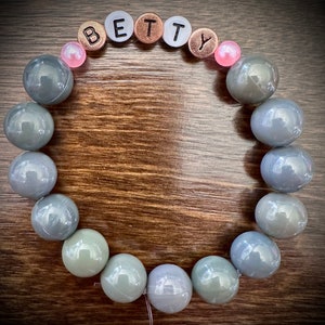 May include: A gray beaded bracelet with the name "BETTY" spelled out in silver and copper letters. The bracelet has pink beads on either side of the letters.