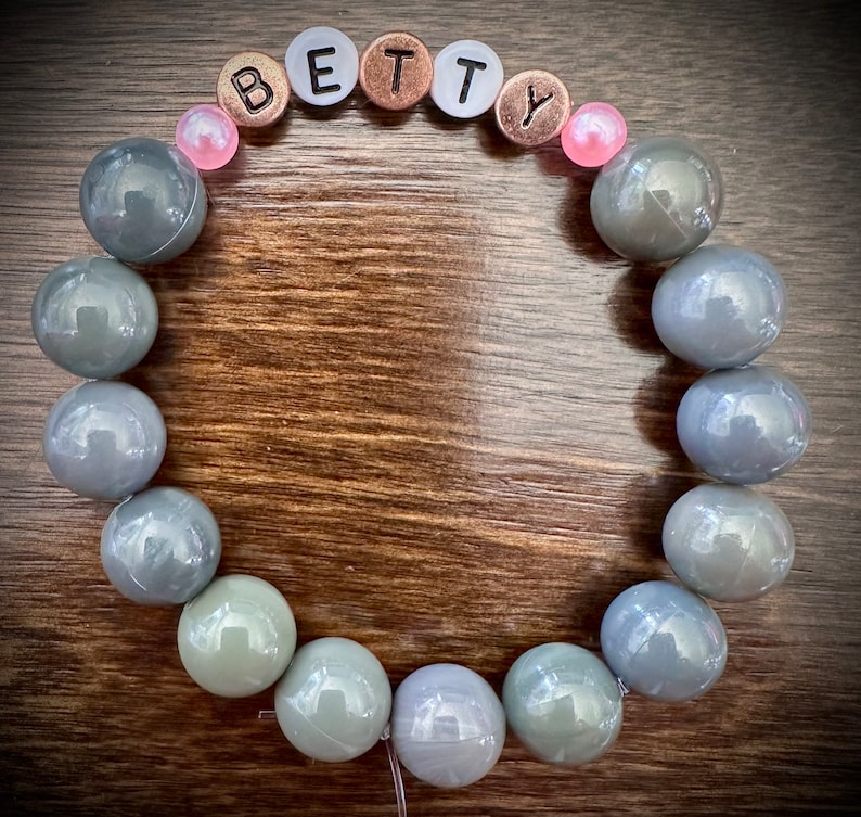 May include: A gray beaded bracelet with the name "BETTY" spelled out in silver and copper letters. The bracelet has pink beads on either side of the letters.
