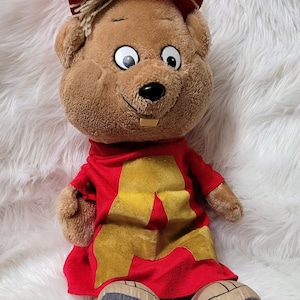 May include: A brown plush toy squirrel wearing a red hat with a gold letter 'A' on a red shirt. The squirrel is sitting on a white fluffy surface.