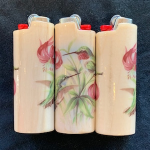 May include: Three wooden lighters with a floral design featuring hummingbirds and pink lilies. The lighters are cream colored with a wood grain texture.