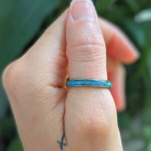 May include: A turquoise and brown ring is worn on a finger. The ring has a thin band with a textured, colorful design. The background is a soft green, suggesting an outdoor setting. The ring's design is the focal point.