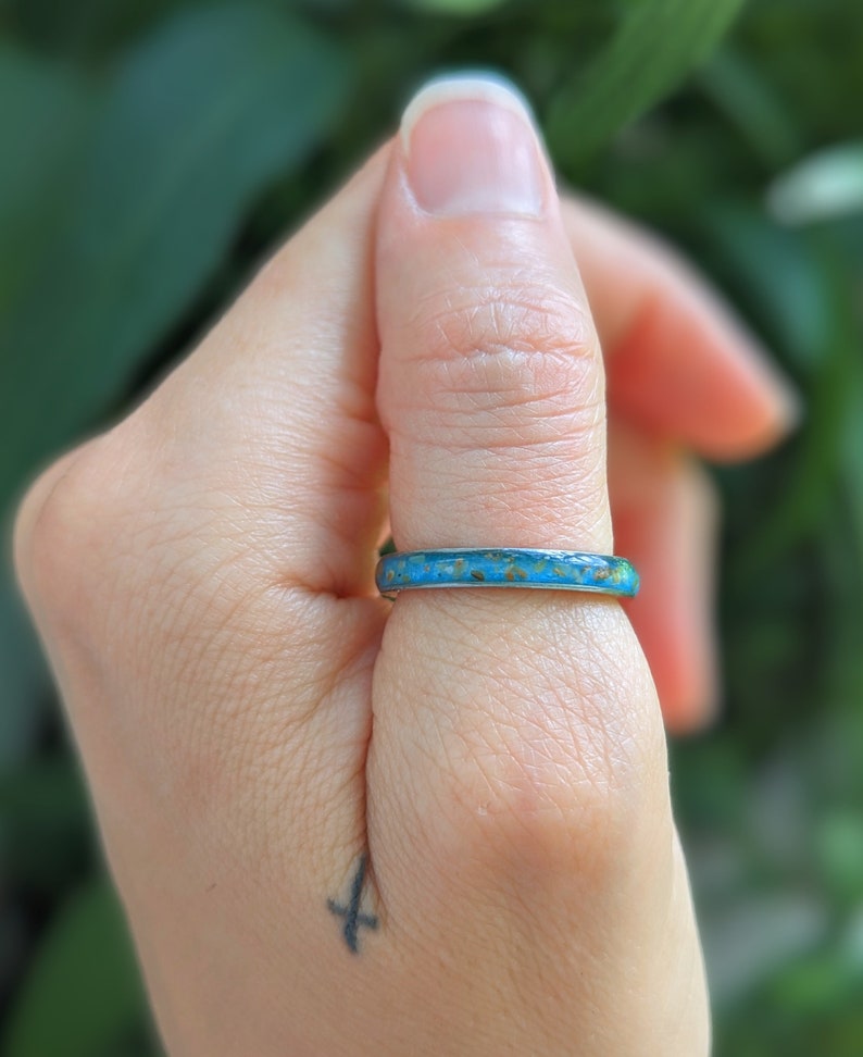 May include: A turquoise and brown ring is worn on a finger. The ring has a thin band with a textured, colorful design. The background is a soft green, suggesting an outdoor setting. The ring's design is the focal point.