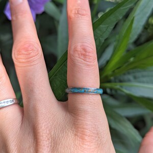 May include: A silver ring with a turquoise and brown inlay is worn on a finger. The ring is set against a backdrop of green leaves and a purple flower, creating a natural aesthetic.