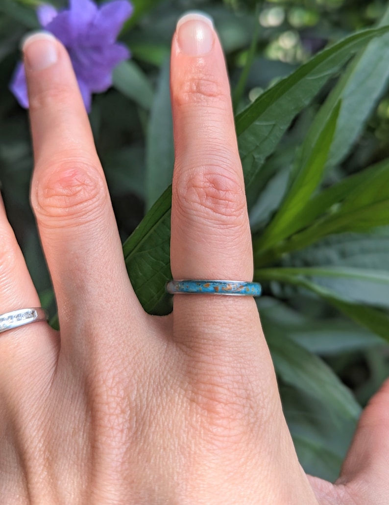 May include: A silver ring with a turquoise and brown inlay is worn on a finger. The ring is set against a backdrop of green leaves and a purple flower, creating a natural aesthetic.
