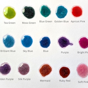 May include: A collection of colorful paint swatches on a white background. The swatches include shades of green, blue, purple, and pink, with names like "Tea Green," "Moss Green," and "Ruby Red" written below each. The paint appears glossy and textured.