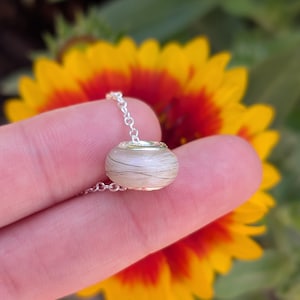 May include: A small, round pendant necklace with a silver chain. The pendant is a clear, glass-like bead with strands of hair encased inside. The background is a blurred image of a yellow and red flower.