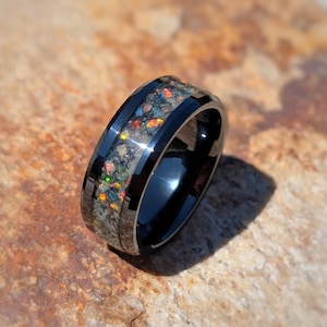 May include: A black ring with a band of colorful, iridescent material. The ring has a polished black finish with a contrasting band of textured, speckled material. The ring is set against a textured, brown surface.